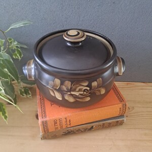 May include: A dark brown ceramic pot with a matching lid, featuring two side handles and a hand-painted beige floral motif on the front. The lid has a small, circular knob with concentric rings in brown and beige tones, and the pot has a smooth finish.