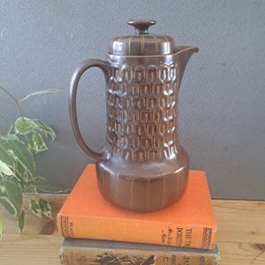 May include: A brown ceramic coffee pot with a lid, featuring a textured, repeating arch pattern on the upper body and vertical stripes on the base. The pot has a curved handle and a spout, showcasing a glossy finish throughout the entire piece.