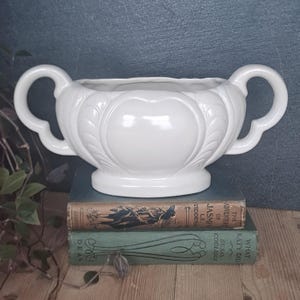 May include: A white ceramic planter with two curved handles and a heart-shaped design. The planter is on two vintage books. The background is a dark teal colour. The books have gold lettering.