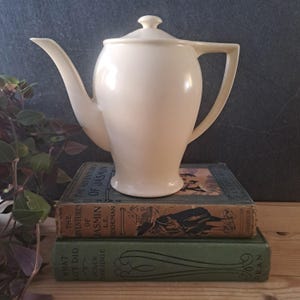 May include: A cream-coloured ceramic teapot with a long spout and handle. It rests on a stack of vintage books, including "The Adventures of Jasmin". The background is a dark grey, with a hint of foliage.