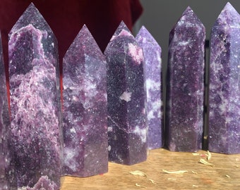 May include: A collection of eight purple and white lepidolite crystal points, arranged in a row on a wooden surface. The crystals are all roughly the same size and shape, and they have a natural, unpolished finish.