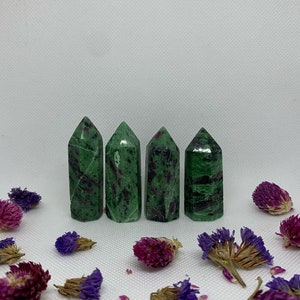 Puede incluir: Cuatro puntas de cristal de zultanita verde y rosa dispuestas en fila sobre una superficie blanca con flores moradas y marrones esparcidas alrededor.