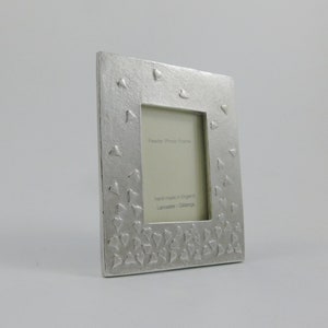 May include: A rectangular pewter photo frame with a textured surface and heart-shaped embellishments. The frame has a central opening for a photo and includes the text "Pewter Photo Frame" and "hand made in England Lancaster Gibbins".