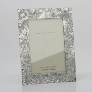 May include: A rectangular pewter photo frame with a textured, metallic silver finish featuring an abstract pattern of intersecting lines and small floral motifs. The insert reads Pewter Photo Frame 6 x 4 inches, hand made in England, Lancaster & Gibbings.