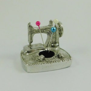 May include: A silver sewing machine pin cushion with three pins. The pin cushion is shaped like a vintage sewing machine with decorative details. The pins have pink, blue, and white heads. Two silver thread spools are on the base.