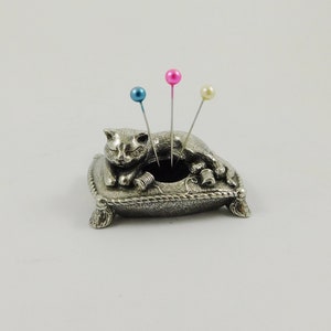 May include: A silver-coloured cat pin cushion with three pins. The cat is lying on a pillow with a decorative rope trim. The pins have blue, pink, and cream-coloured heads. The cushion has a small opening for storing pins.