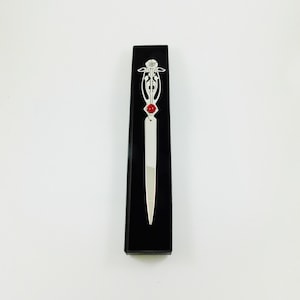May include: A silver letter opener with an Art Nouveau design and a red gemstone accent, presented in a black box. The decorative top features floral and flowing lines.