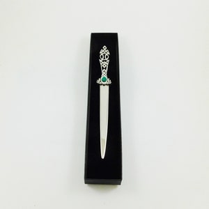 May include: A silver letter opener with an ornate handle and a green gemstone, presented in a black box. The handle features Celtic knotwork. The blade is long and slender, and the overall design is elegant.