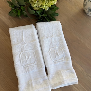 May include: Two white rectangular cotton towels featuring a textured terry cloth finish. Each towel displays a centered, embroidered square emblem containing a single capital letter, with one towel featuring the letter M and the other the letter A.