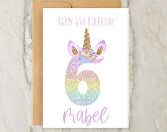 May include: A white card with a unicorn-themed design. The card says "Happy 6th Birthday" and features a rainbow glitter number 6 with a unicorn horn and ears. The name "Mabel" is written in purple below the number.