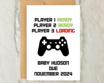 Op de afbeelding: Een witte kaart met een zwarte afbeelding van een videogamecontroller en de tekst "PLAYER 1 READY, PLAYER 2 READY, PLAYER 3 LOADING". Onder de controller staat de tekst "BABY HUDSON DUE NOVEMBER 2024".