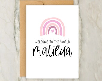 May include: A white card with a pink and purple rainbow with a small pink heart in the center. The text "WELCOME TO THE WORLD matilda" is printed below the rainbow.