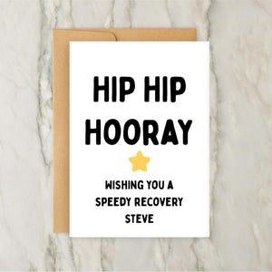 Peut inclure: Une carte de vœux blanche avec l'inscription "HIP HIP HOORAY" en noir, une étoile jaune et le message "WISHING YOU A SPEEDY RECOVERY STEVE". La carte est dans une enveloppe marron.