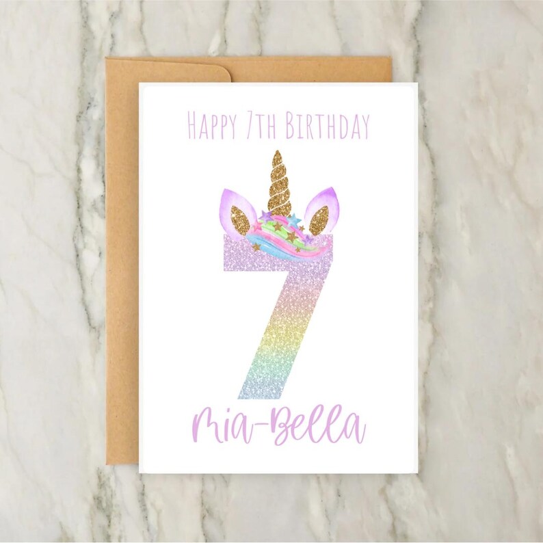 Puede incluir: Una tarjeta blanca con una cabeza de unicornio y el n&uacute;mero 7 en purpurina arco&iacute;ris. El texto "Happy 7th Birthday" y "Mia-Bella" est&aacute;n impresos en morado.