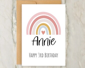 Op de afbeelding: Een witte kaart met een roze, gele en blauwe regenboog met een klein hartje in het midden. De naam "Annie" staat in zwarte cursieve letters onder de regenboog. De tekst "Happy 3rd Birthday" staat in het zwart onder de naam.