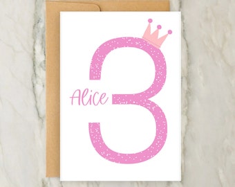Op de afbeelding: Een witte wenskaart met een groot roze cijfer drie met een wit spikkelpatroon en een kleine roze kroon aan de bovenkant. De naam Alice is in roze schrijfletters links van het cijfer gedrukt. Een bruine envelop zit achter de kaart.