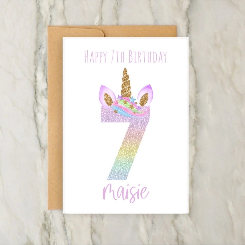 Puede incluir: Una tarjeta blanca con una cabeza de unicornio de purpurina arco&iacute;ris y el n&uacute;mero 7. El texto "Happy 7th Birthday Maisie" est&aacute; impreso en la tarjeta.