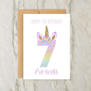 Puede incluir: Una tarjeta blanca con una cabeza de unicornio y el n&uacute;mero 7 en purpurina arco&iacute;ris. El texto "Happy 7th Birthday Miranda" est&aacute; impreso en la tarjeta.