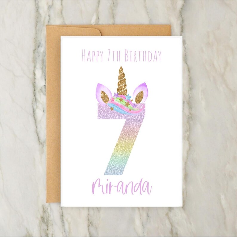 Puede incluir: Una tarjeta blanca con una cabeza de unicornio y el n&uacute;mero 7 en purpurina arco&iacute;ris. El texto "Happy 7th Birthday Miranda" est&aacute; impreso en la tarjeta.