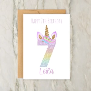 Puede incluir: Una tarjeta blanca con una cabeza de unicornio arco&iacute;ris brillante y el n&uacute;mero 7. El texto "Happy 7th Birthday Leila" est&aacute; impreso en la tarjeta.