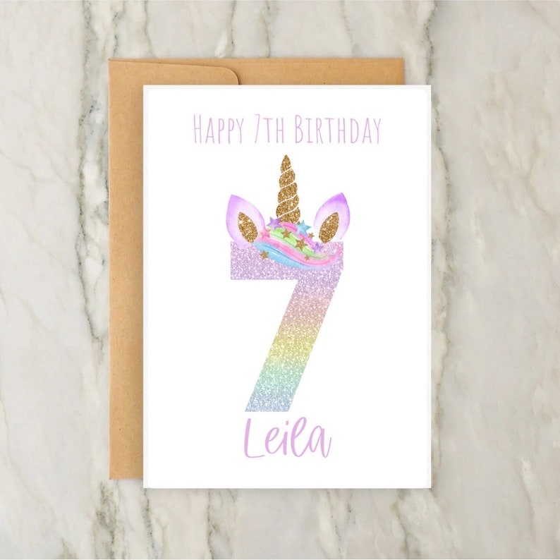 Puede incluir: Una tarjeta blanca con una cabeza de unicornio arco&iacute;ris brillante y el n&uacute;mero 7. El texto "Happy 7th Birthday Leila" est&aacute; impreso en la tarjeta.