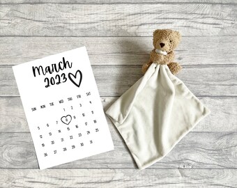 May include: A white paper calendar for March 2023 with a heart icon around the number 8, positioned next to a light beige plush teddy bear security blanket featuring a small stuffed bear head attached to a soft, square-shaped fabric cloth.