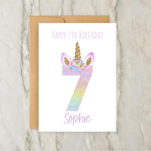 Puede incluir: Una tarjeta blanca con un dise&ntilde;o de unicornio para un 7&ordm; cumplea&ntilde;os. La tarjeta presenta un n&uacute;mero 7 brillante con orejas y un cuerno de unicornio. El texto "Happy 7th Birthday" y "Sophie" est&aacute;n impresos en la tarjeta.