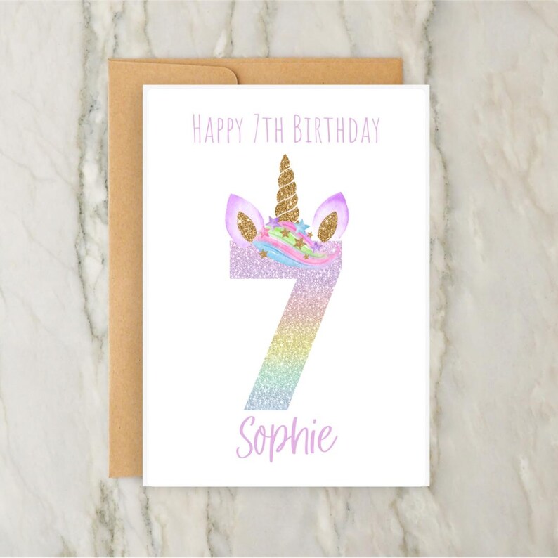 Puede incluir: Una tarjeta blanca con un dise&ntilde;o de unicornio para un 7&ordm; cumplea&ntilde;os. La tarjeta presenta un n&uacute;mero 7 brillante con orejas y un cuerno de unicornio. El texto "Happy 7th Birthday" y "Sophie" est&aacute;n impresos en la tarjeta.