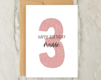 Op de afbeelding: Een witte verjaardagskaart met een groot, oudroze cijfer drie met een wit spikkelpatroon. De tekst luidt Happy Birthday in een eenvoudig zwart lettertype, gevolgd door de naam Maddie in een zwart cursief handschrift, met een bruine envelop.