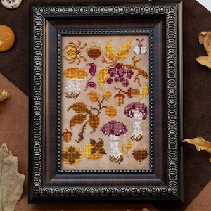 May include: A framed cross-stitch with an autumnal design, including mushrooms, leaves, and berries in brown, yellow, and purple hues. The dark brown frame is surrounded by dried leaves and decorative buttons, evoking a seasonal theme.
