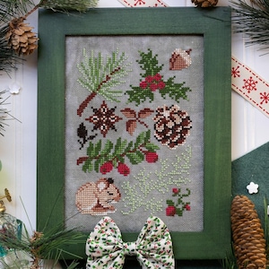 May include: A framed cross-stitch featuring botanical elements like pine needles, berries, and a pinecone. The frame is green, and the artwork is surrounded by pine branches, a decorative bow, and a green glass lamp. A festive ribbon with snowflakes adds to the holiday theme.
