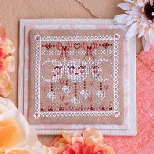 Puede incluir: Un diseño de punto de cruz con un tema celestial, con lunas, corazones y detalles florales en tonos blancos, rosas y burdeos. El diseño está sobre una tela beige, rodeado de arreglos florales y un fondo rosa.