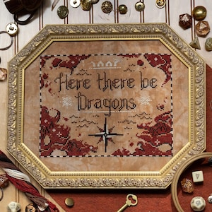 Puede incluir: Obra de arte de punto de cruz en un marco dorado adornado, con el texto "Here there be Dragons" con una rosa de los vientos y motivos de dragones en rojo y negro. El marco está rodeado de nociones de costura, dados y una llave.