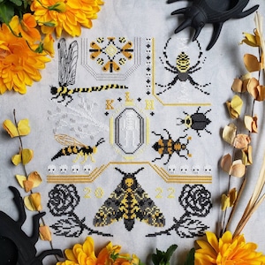 A finished cross stitch featuring two large rose motifs flanking a Deaths-head hawkmoth, a large wasp, a dragonfly, beetles, and an orb weaver spider. A simple skeleton is in the middle of the design and various skull motifs are throughout.