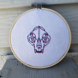 以下が含まれることがあります： 赤と青の3D刺繍で動物の頭蓋骨が刺繍された白い刺繍枠です。