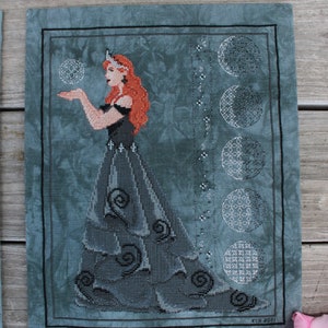 Puede incluir: Un patrón de punto de cruz que presenta a una mujer con un vestido azul y negro con una corona en la cabeza. Ella sostiene una esfera en su mano. El fondo es un degradado azul y blanco con una fila de lunas en diferentes fases.