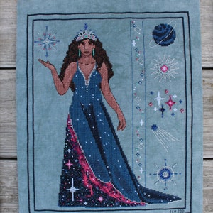 Può includere: Un ricamo a punto croce di una donna che indossa un abito blu con un design di galassia rosa e nero. Indossa una corona e tiene una stella. Lo sfondo è blu chiaro con stelle e pianeti.