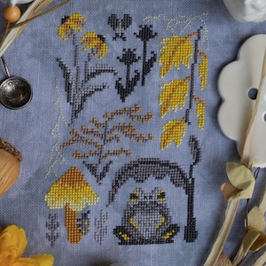 May include: Cross-stitch embroidery on a blue fabric featuring a gray frog sitting under a gray archway, surrounded by yellow flowers, mushrooms, and leaves.