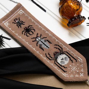 Könnte beinhalten: Beige Kreuzstich-Lesezeichen mit schwarzen und grauen Insekten. Das Lesezeichen hat eine Perlenquaste und eine silberne Kette. Eine bernsteinfarbene Glasflasche und schwarze Spinnen sind im Hintergrund.