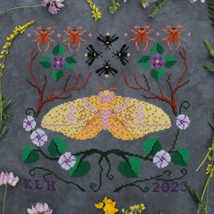 May include: Cross-stitch embroidery of a yellow and pink moth with a brown, green, and purple floral border. The design includes a circle of stylized fireflies and the text "KLN 2023".