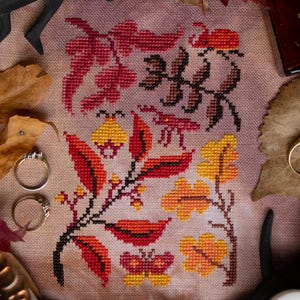 May include: Cross-stitch embroidery featuring a floral design with red, orange, and yellow leaves, a butterfly, and a ladybug. The design is on a beige fabric background.