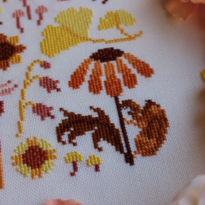 Peut inclure: Broderie au point de croix repr&eacute;sentant une vari&eacute;t&eacute; de fleurs et de feuilles d'automne dans des tons d'orange, de jaune et de brun. Le motif comprend des tournesols, des marguerites et des feuilles.