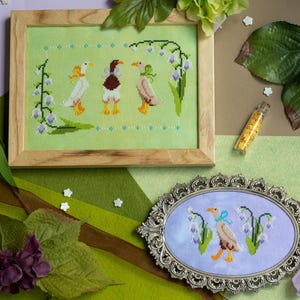 Puede incluir: Un bordado a punto de cruz enmarcado con tres patos y detalles florales. Los patos tienen patas y picos amarillos y son de varios colores. También hay un bordado a punto de cruz ovalado con un pato y flores.