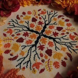 May include: A cross-stitch embroidery of a tree with a circular design of autumn leaves in shades of orange, yellow, and red. The tree branches are black and the background is a light beige.