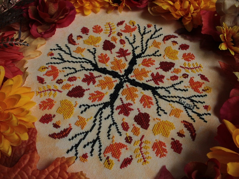 May include: A cross-stitch embroidery of a tree with a circular design of autumn leaves in shades of orange, yellow, and red. The tree branches are black and the background is a light beige.