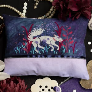 May include: A decorative pillow with a cross-stitched design featuring a white creature with purple spots, set against a dark blue background with red and blue floral elements. The bottom half is a light purple. A black ribbon accents the design.