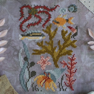 Könnte beinhalten: Kreuzstich-Stickerei mit einem farbenfrohen Unterwassermotiv mit einem Oktopus, Fischen, Algen und Korallen. Das Design ist auf einem hellgrauen Stoff gestickt.