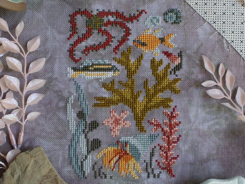 K&ouml;nnte beinhalten: Kreuzstich-Stickerei mit einem farbenfrohen Unterwassermotiv mit einem Oktopus, Fischen, Algen und Korallen. Das Design ist auf einem hellgrauen Stoff gestickt.