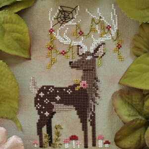 May include: Cross-stitch design of a deer with white antlers adorned with flowers and a spiderweb. The deer is brown with white spots and stands in front of mushrooms and greenery. The background is a neutral tan color.