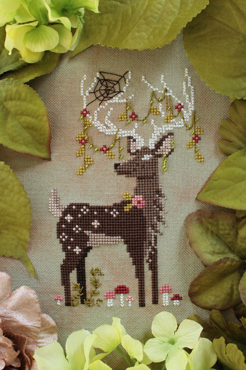May include: Cross-stitch design of a deer with white antlers adorned with flowers and a spiderweb. The deer is brown with white spots and stands in front of mushrooms and greenery. The background is a neutral tan color.